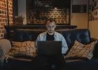 Man in black crew neck t shirt and denim jacket sitting on a couch using his laptop